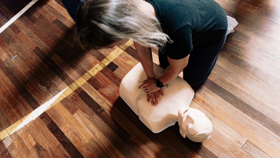 Person performing chest compressions on a CPR manikin on a wooden gym floor, training scene