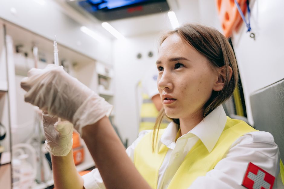 Young woman in a yellow safety vest wearing gloves, inspecting a syringe in a clinical setting.